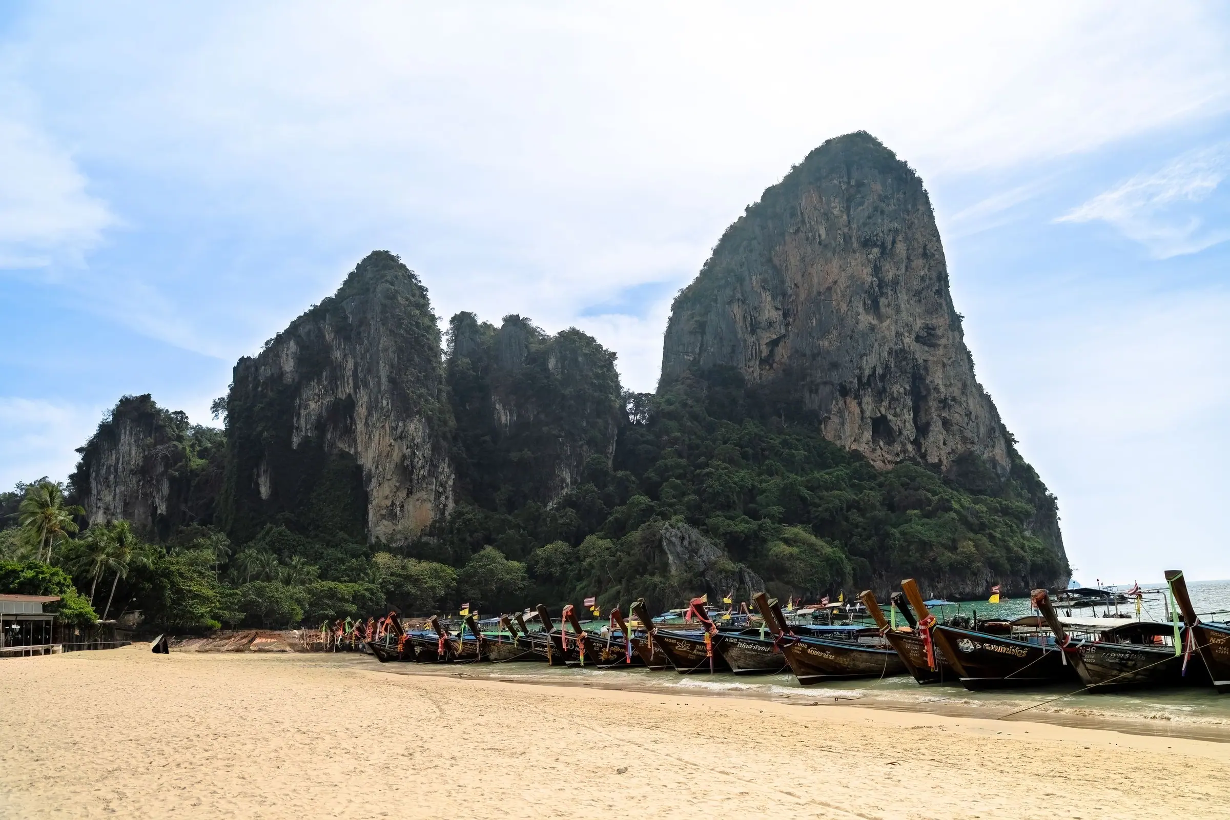 Towering limestone cliffs and long-tail boats at Railay Beach in Thailand with tourists on the shore.